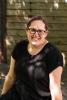 Jennifer sits outside in a black dress in front of a wooden wall in the shade.