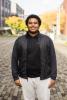 Jamal stands on a gray brick walkway with college buildings behind him.
