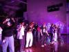 Students dance on a basketball court under pink lighting. A mural made of glow tape is on a concrete wall in the background.