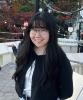 Portrait of a person with long dark hair and glasses standing on an outdoor terrace, with string lights and autumn-colored trees in the background