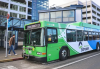 Photo of transit city bus on street in front of city buildings with man in brown jacket and blue jeans standing at bus stop